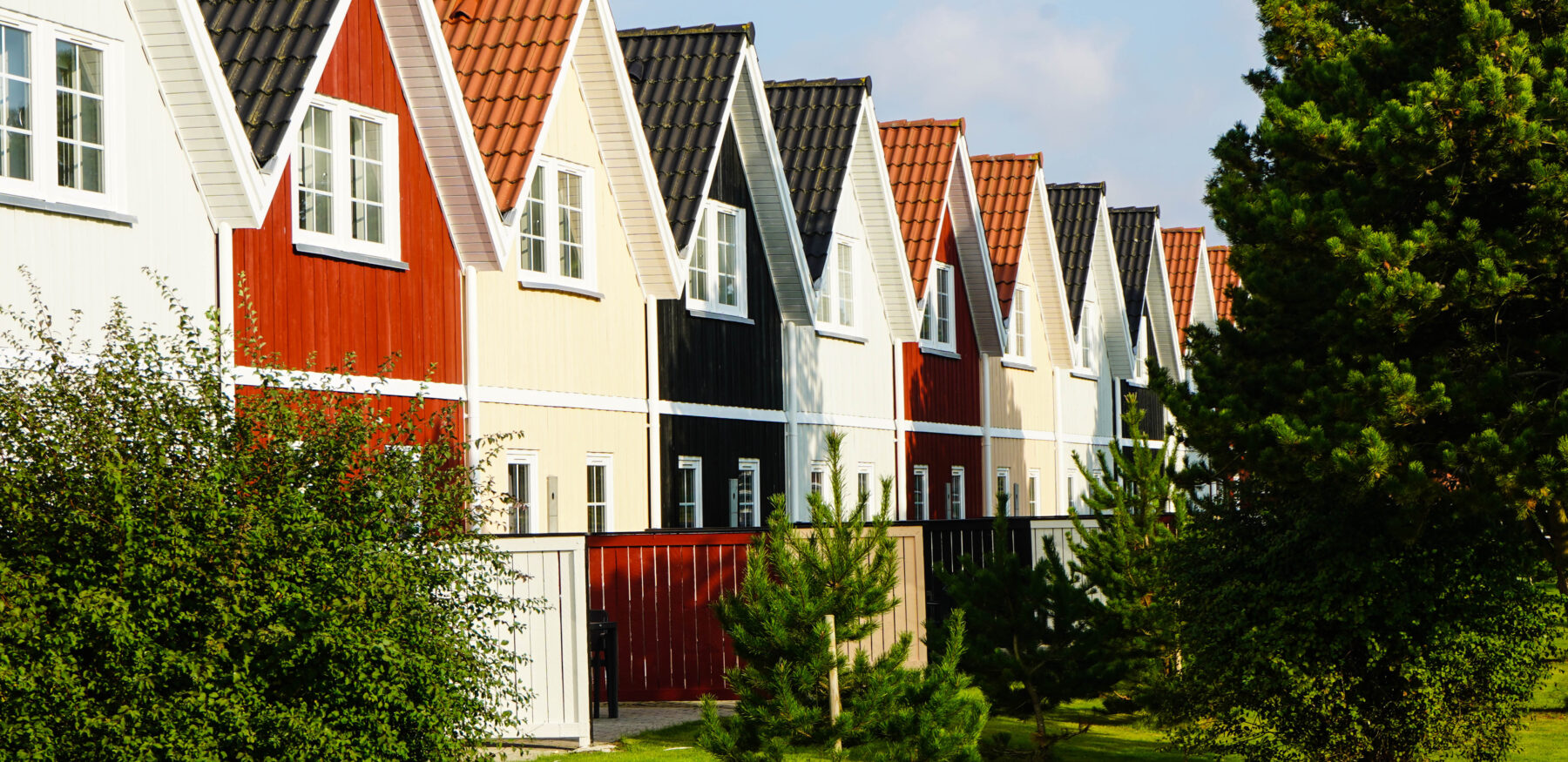 Row of colourful Scandinavian-style holiday homes with steep gabled roofs in Varde Kommune, Denmark, surrounded by lush greenery.