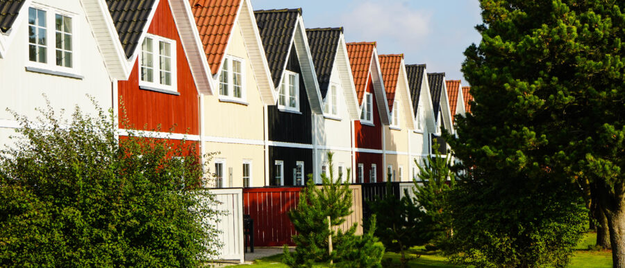 Row of colourful Scandinavian-style holiday homes with steep gabled roofs in Varde Kommune, Denmark, surrounded by lush greenery.