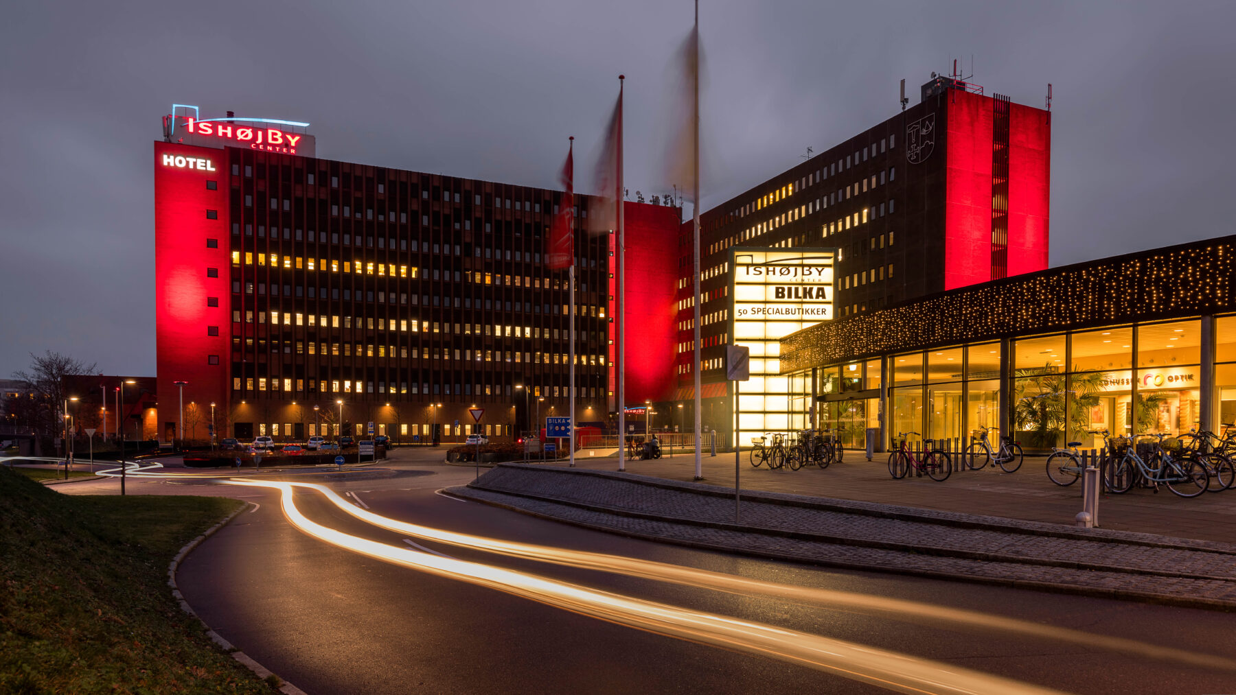 Evening view of Ishøj Shopping Centre with illuminated hotel and Bilka signs, vibrant red lighting, and bicycle parking area in the foreground.