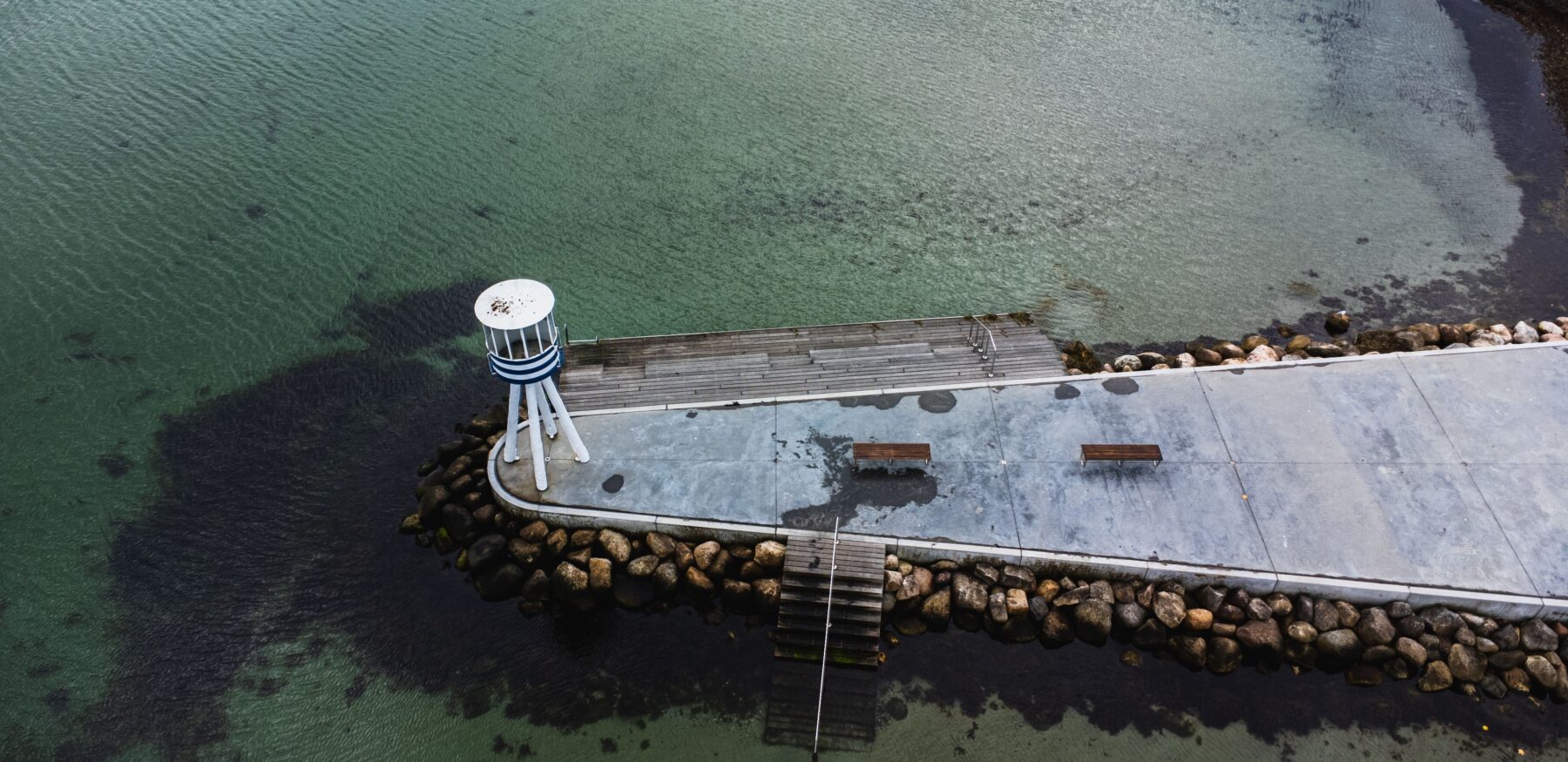 Aerial view of Bellevue Strand pier with clear turquoise water and a small white and blue lifeguard tower, benches, and rocky shoreline.