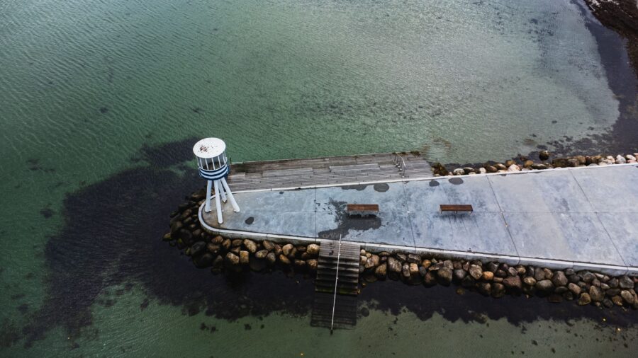 Aerial view of Bellevue Strand pier with clear turquoise water and a small white and blue lifeguard tower, benches, and rocky shoreline.