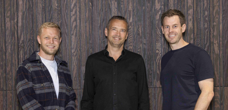 Three members of the Intellis board – Kevin Magnussen, Lars Sneftrup Pedersen and Stig Larsen – standing in front of a dark wooden panel wall, smiling.