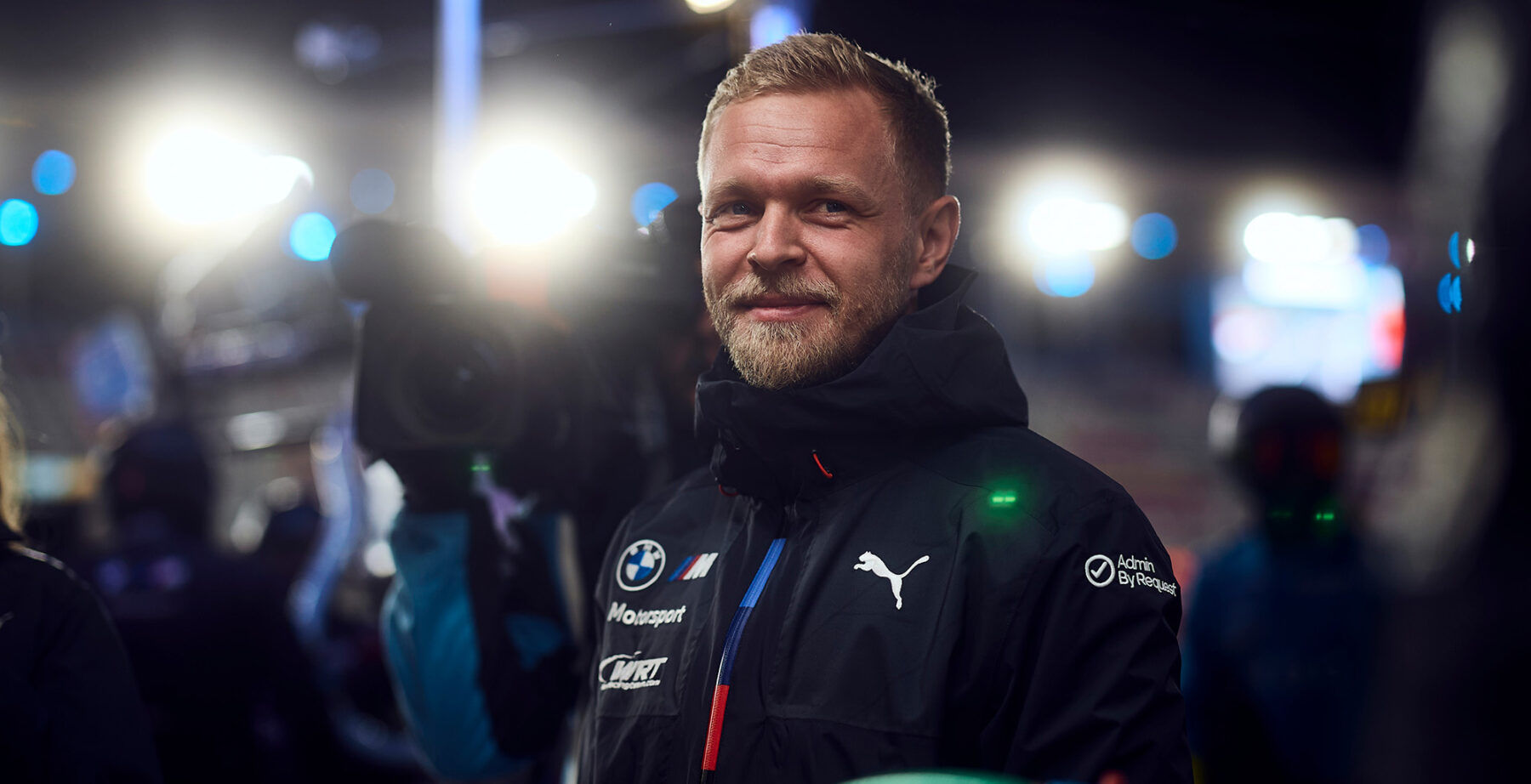 Kevin Magnussen in BMW Motorsport gear, smiling at the camera under bright lights during a night race event.