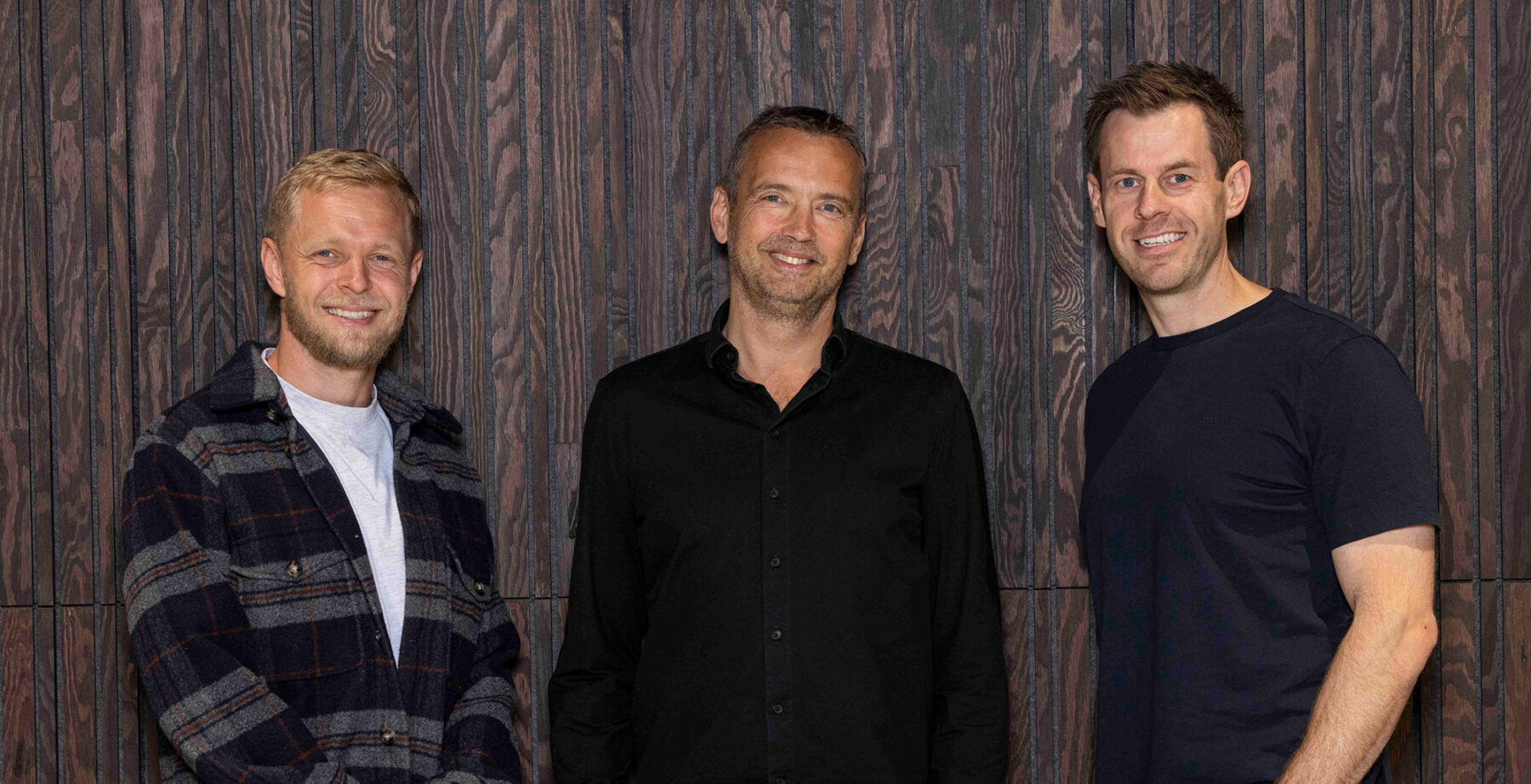 Kevin Magnussen, Lars Sneftrup Pedersen, and Stig Larsen stand smiling in front of a dark wooden wall at Intellis HQ.