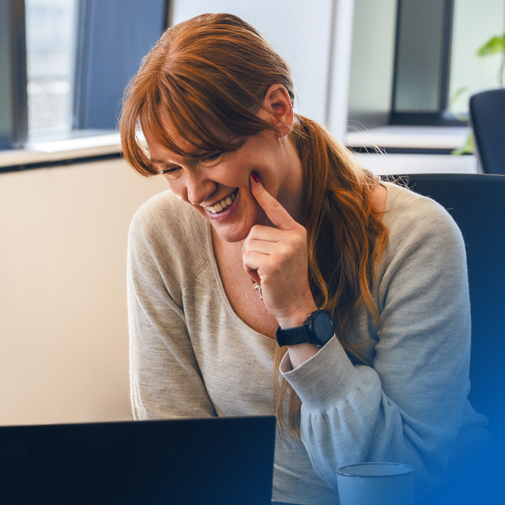 Red-haired professional woman smiling while engaging with her laptop in a modern office environment.