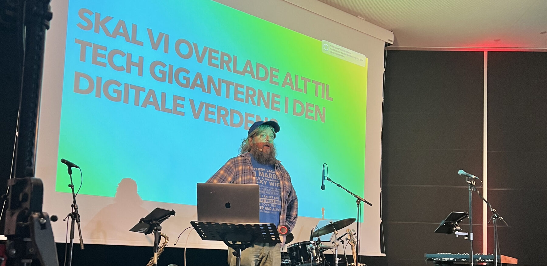 A bearded speaker presents in front of a vibrant slide reading “Skal vi overlade alt til tech giganterne i den digitale verden?”