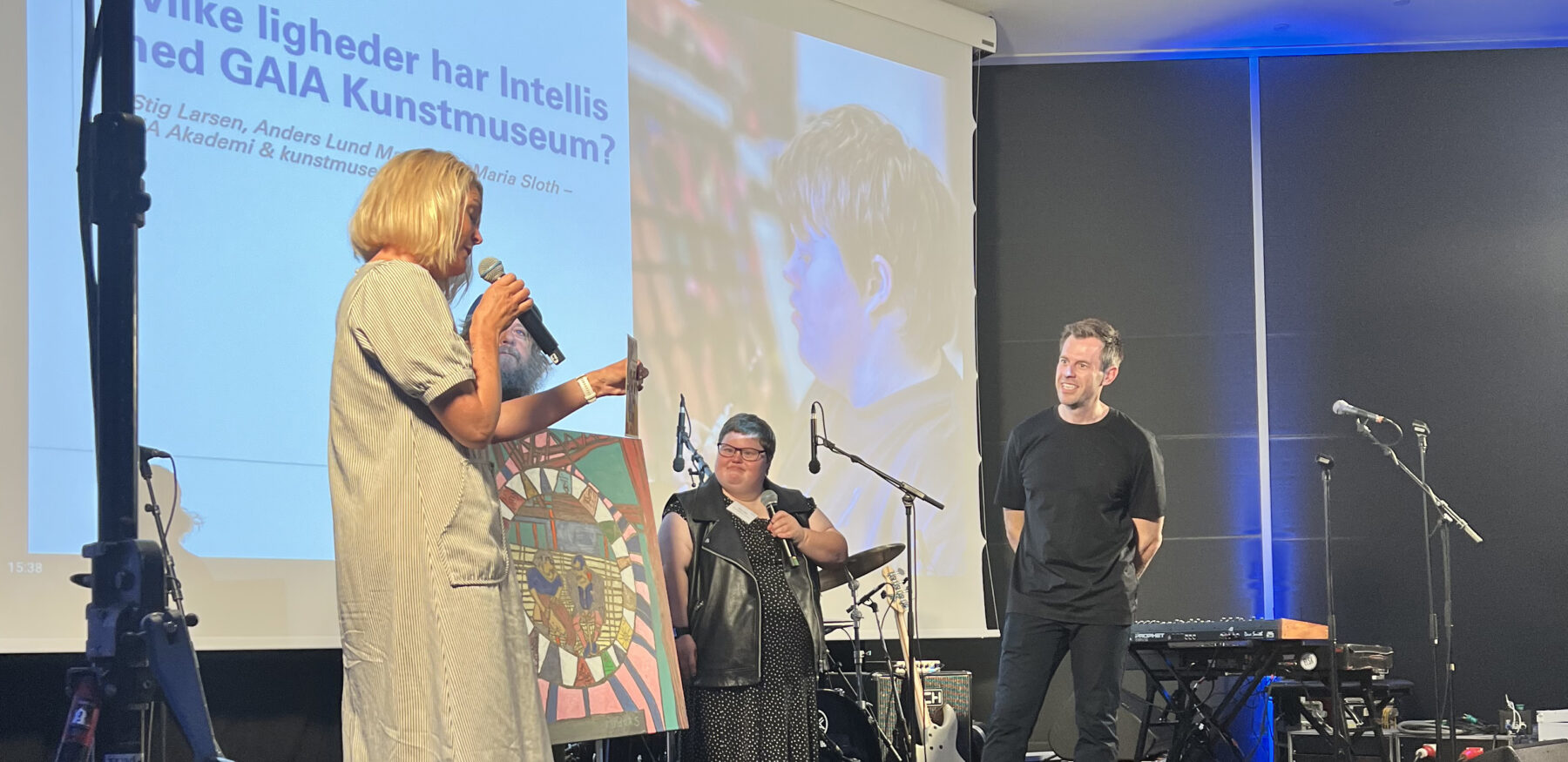 A panel discusses similarities between Intellis and GAIA Art Museum, featuring a speaker, a colourful artwork, and engaged audience members.