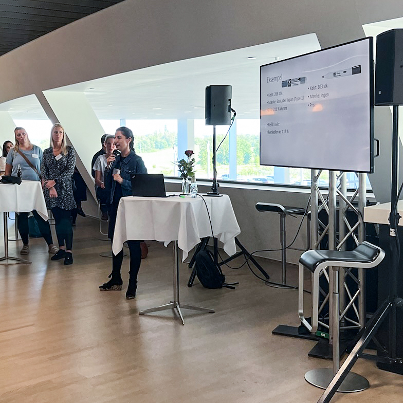 Speaker presenting in a bright, modern conference space with attendees listening at round tables during Intellis Brugerkonference 2023.