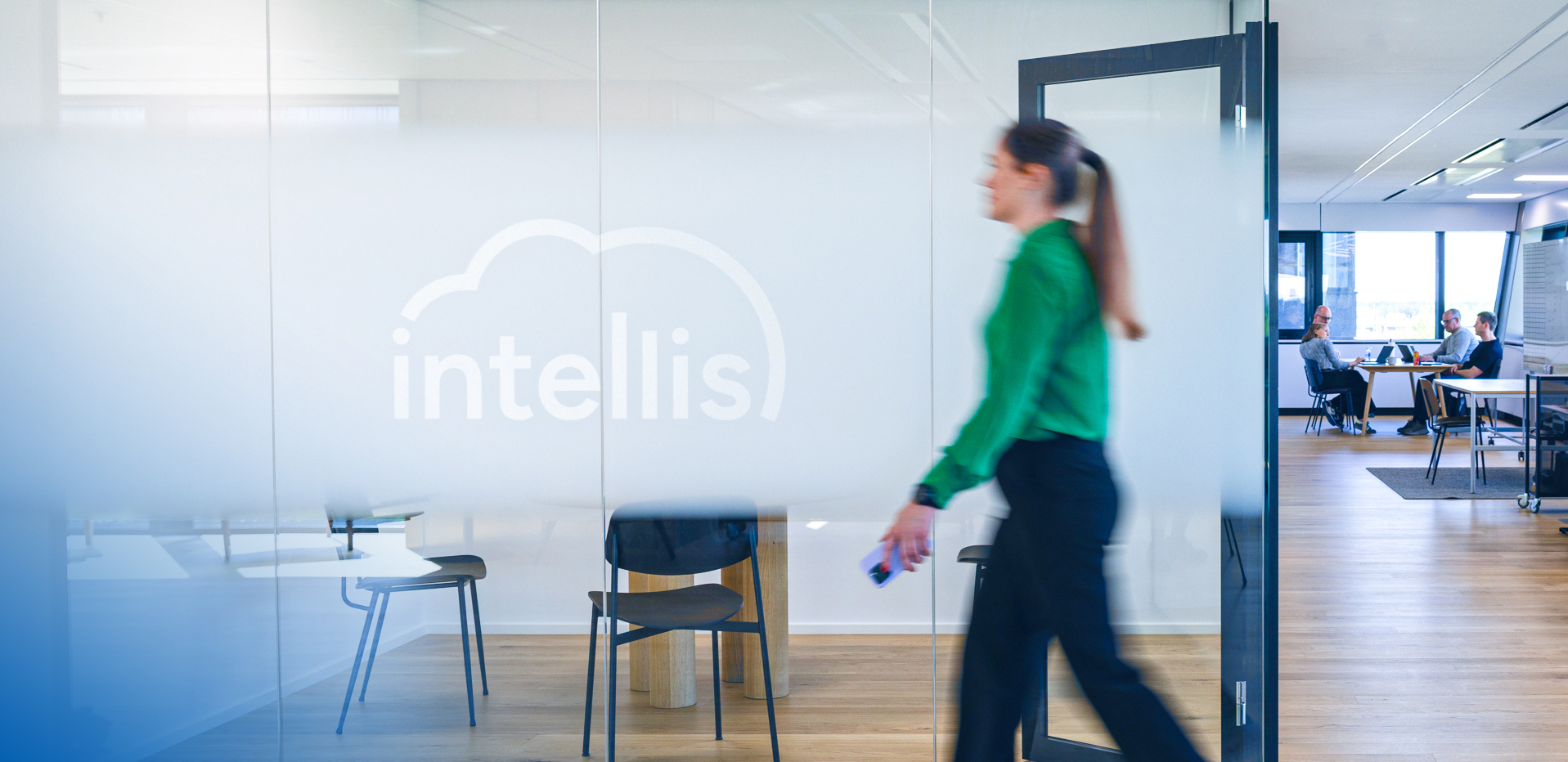 Woman walking past frosted glass wall with Intellis logo at modern office, with team in meeting room in background.