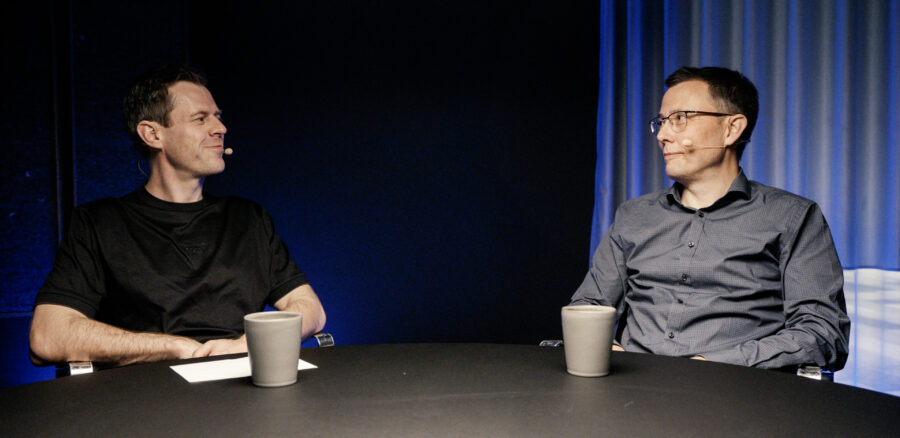 Stig Larsen and Thomas Schultz from Konsido having a business discussion at a round table, both wearing microphones and seated in a modern studio.