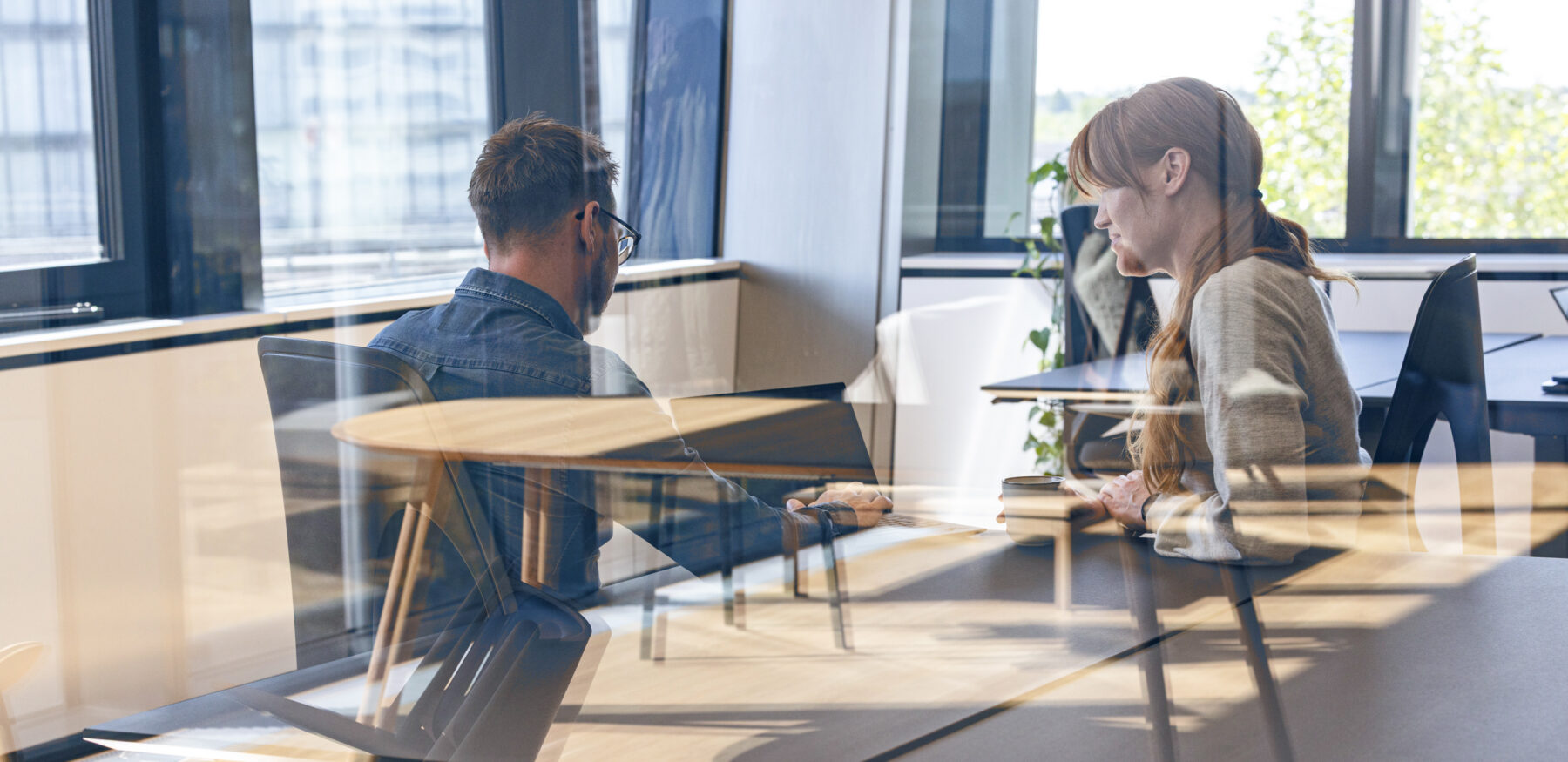 Two colleagues seated at a desk, discussing work on a laptop through a glass-partitioned modern office, with reflections of tables and natural light.