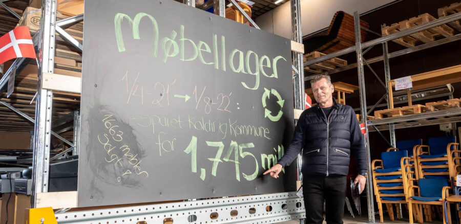 Man standing in warehouse by blackboard sign showing Kolding Municipality’s savings of 1.745 million DKK from reused furniture.