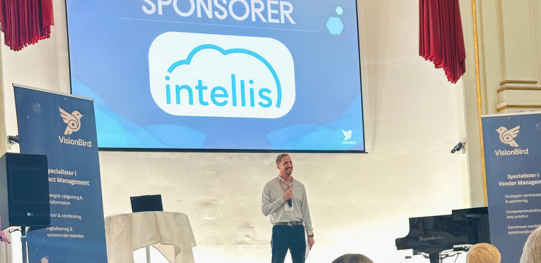 Intellis shown as sponsor on stage at a VisionBird vendor management event with a speaker presenting to an audience.