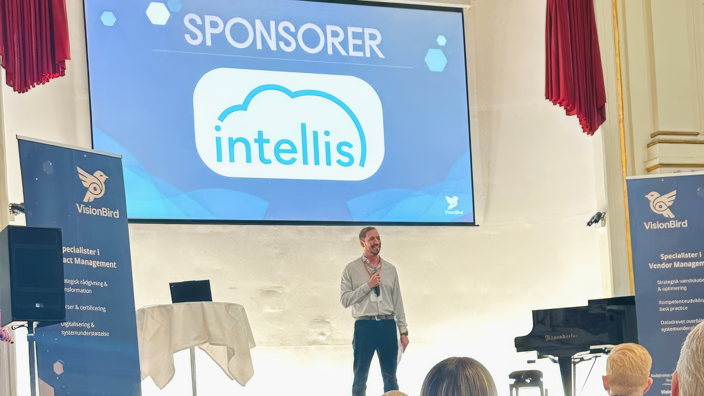 Intellis shown as sponsor on stage at a VisionBird vendor management event with a speaker presenting to an audience.