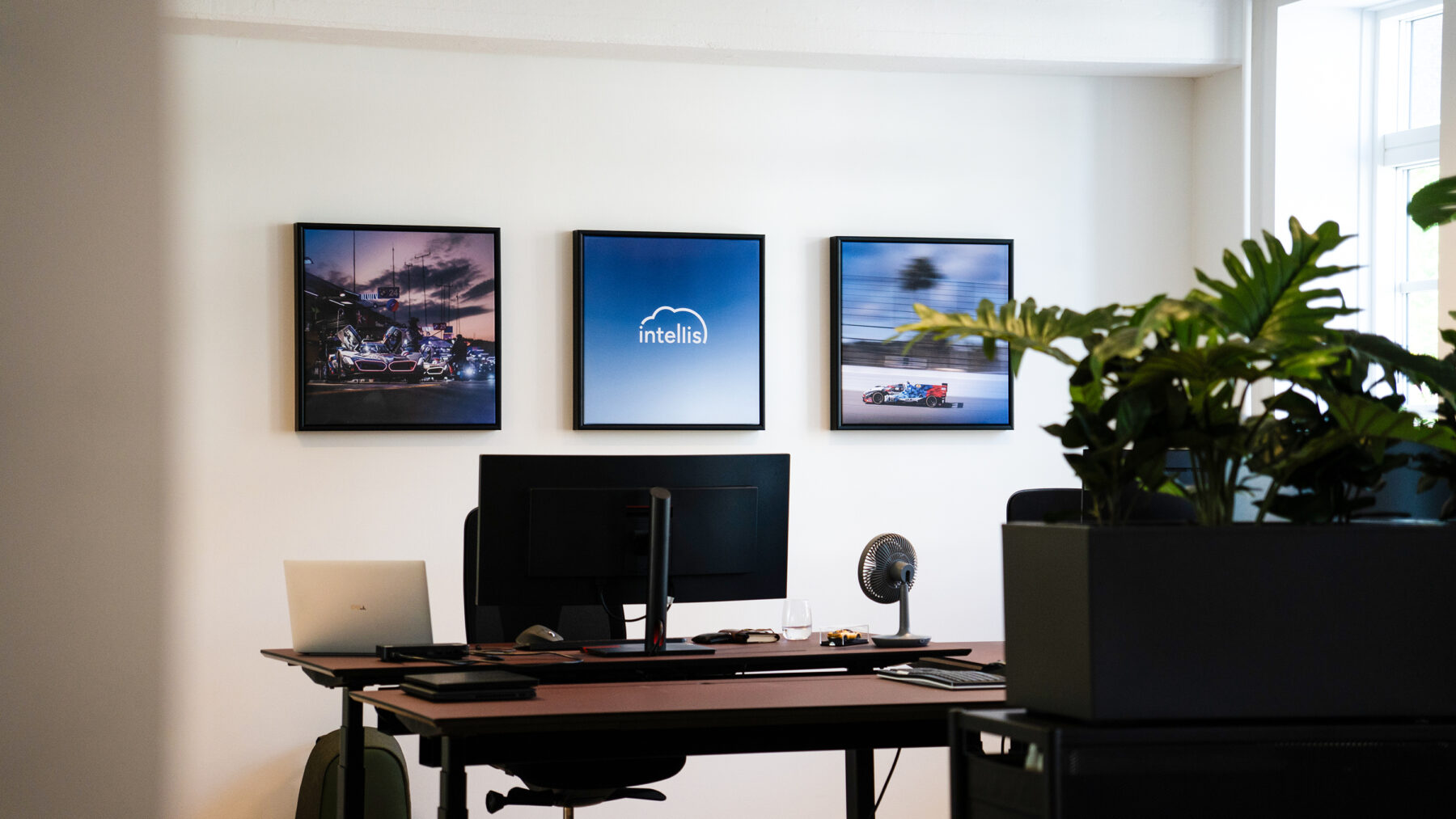 Modern Intellis office workspace with a desk setup, framed racing prints and plants by a bright window.